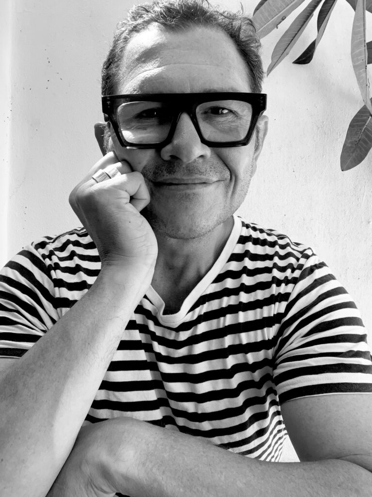 A headshot of Daniel McDonald in black and white. He is wearing glasses and a striped t-shirt while smiling at the camera with his head resting on his hand.