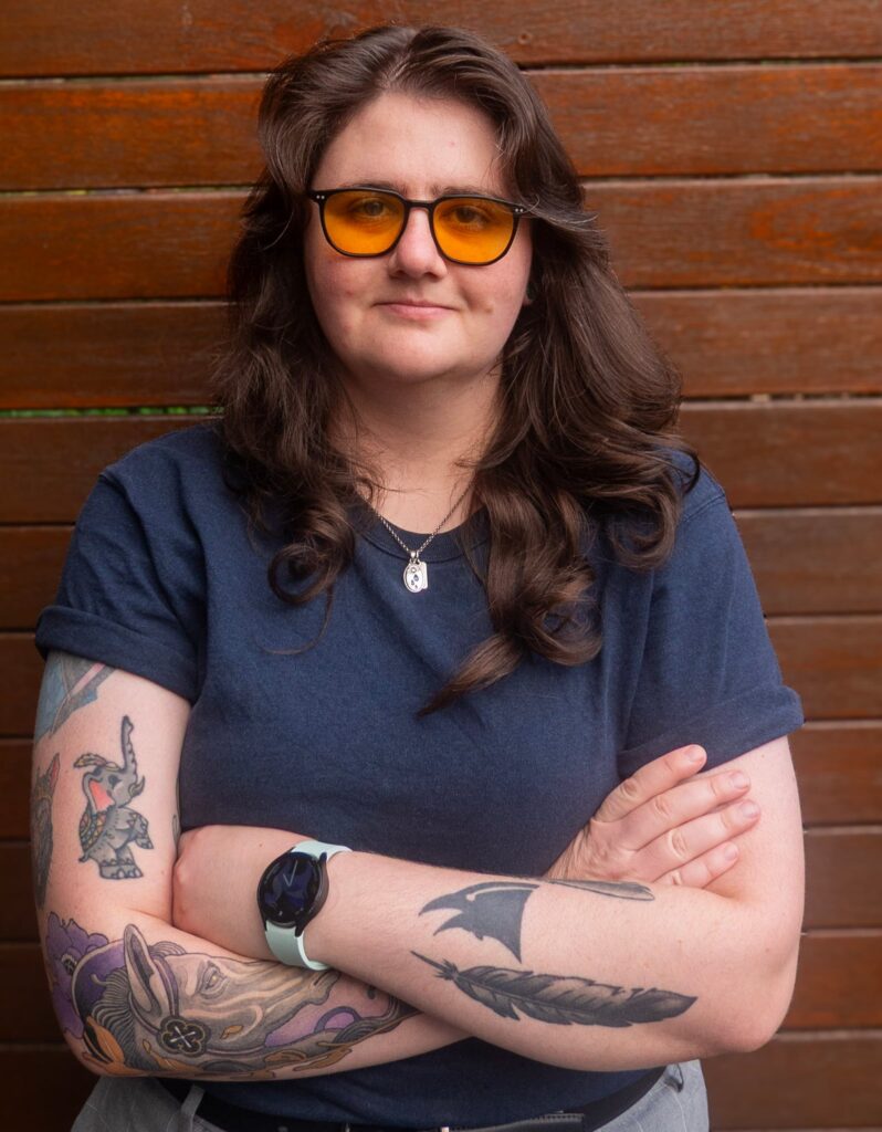 A photo of Dom Parker from the waist up. She is wearing a blue t-shirt and watch on her left wrist with her arms crossed. She has shoulder length brown hair, tattoos on her arms and tinted glasses on her face.