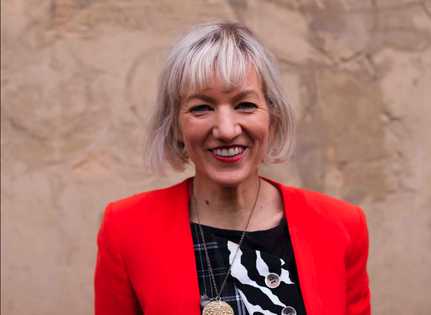 A headshot of ADNA National Director, Sarah-Mace Dennis. She has a short blonde crop and is wearing a red jacket and red lipstick.